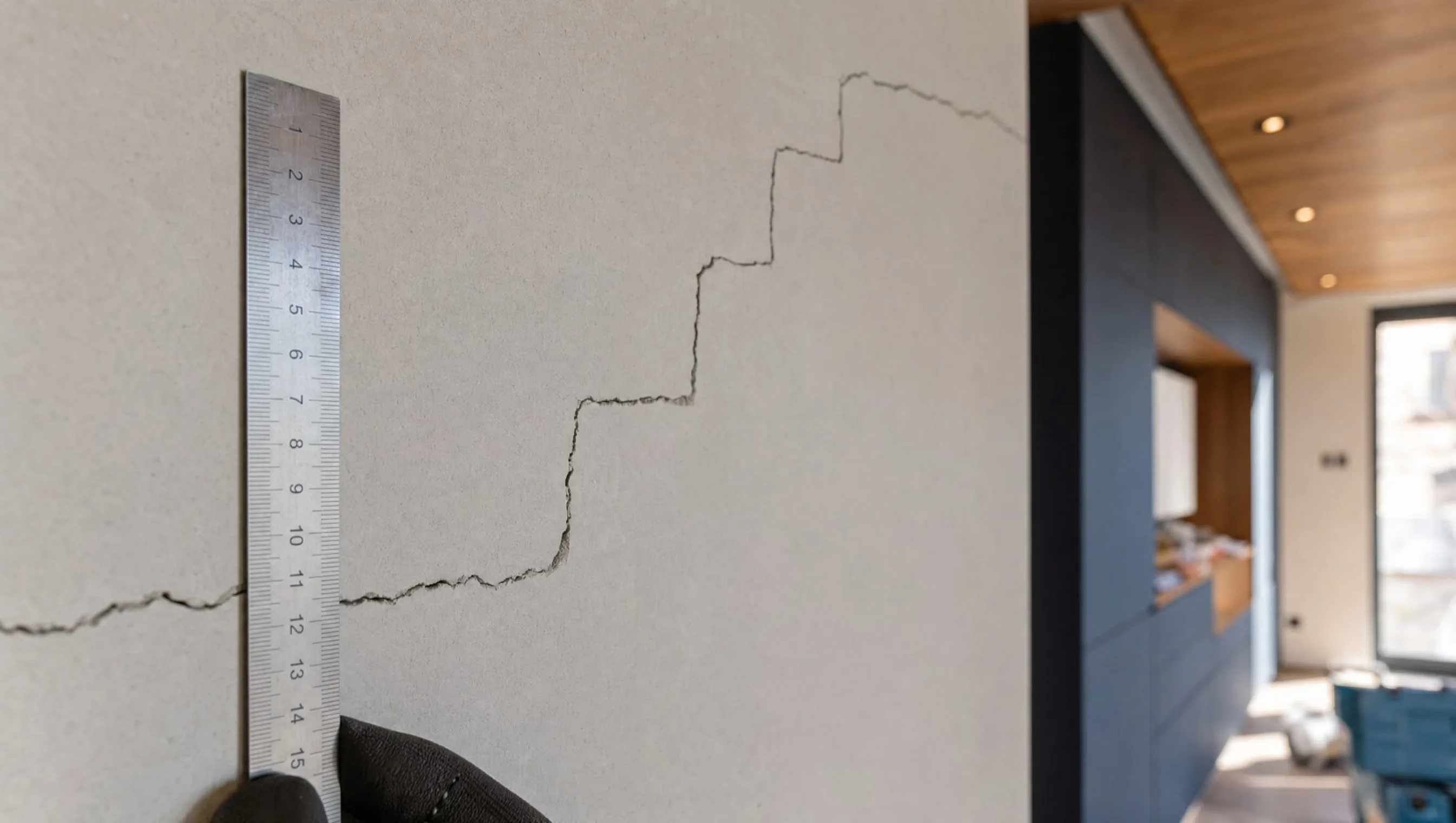 Règle métallique posée à côté d'une fissure murale en escalier, maison neuve Liège, avril 2026